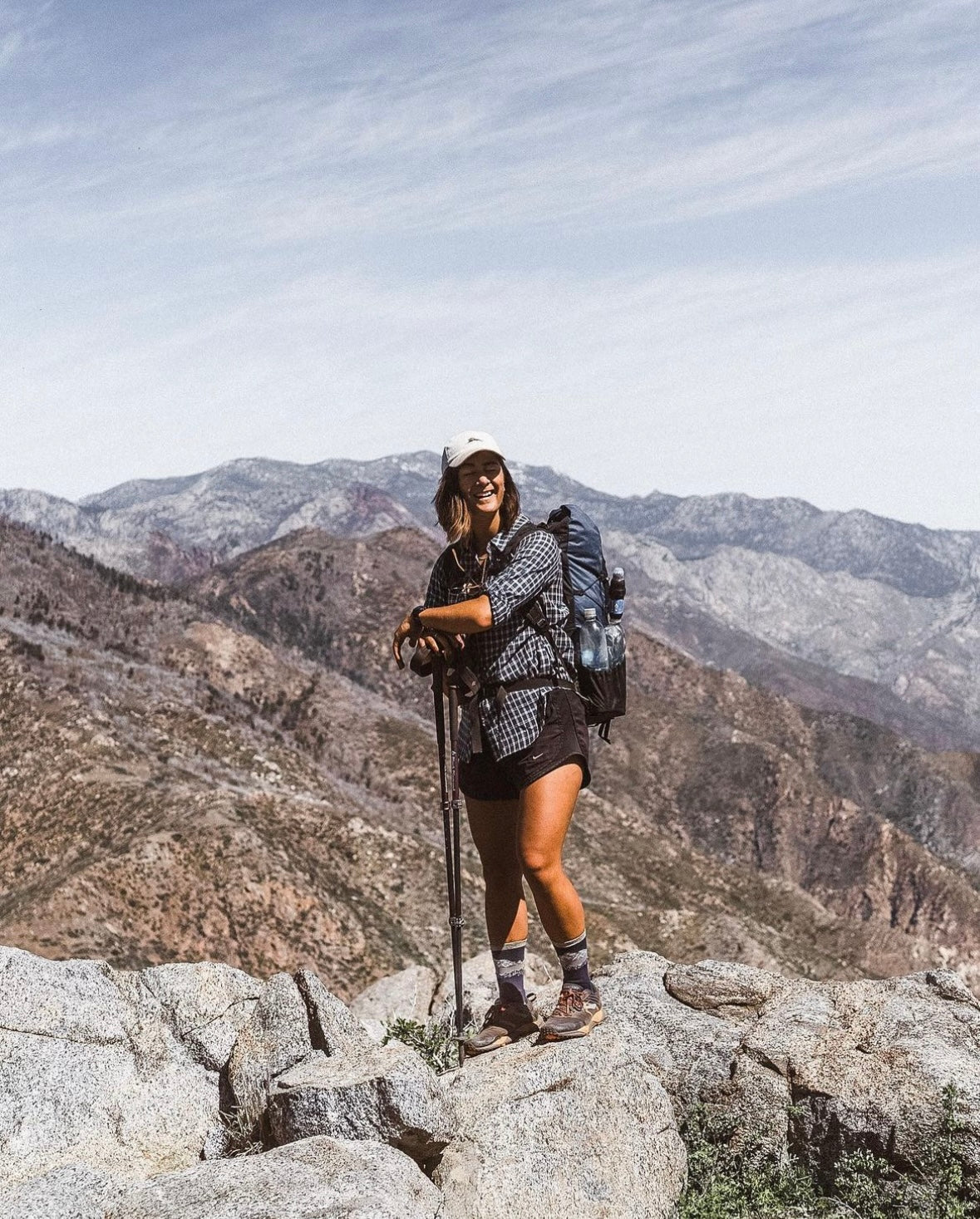 A female hiker smiles at the camera, standing on top of a boulder and resting on her hiking poles.