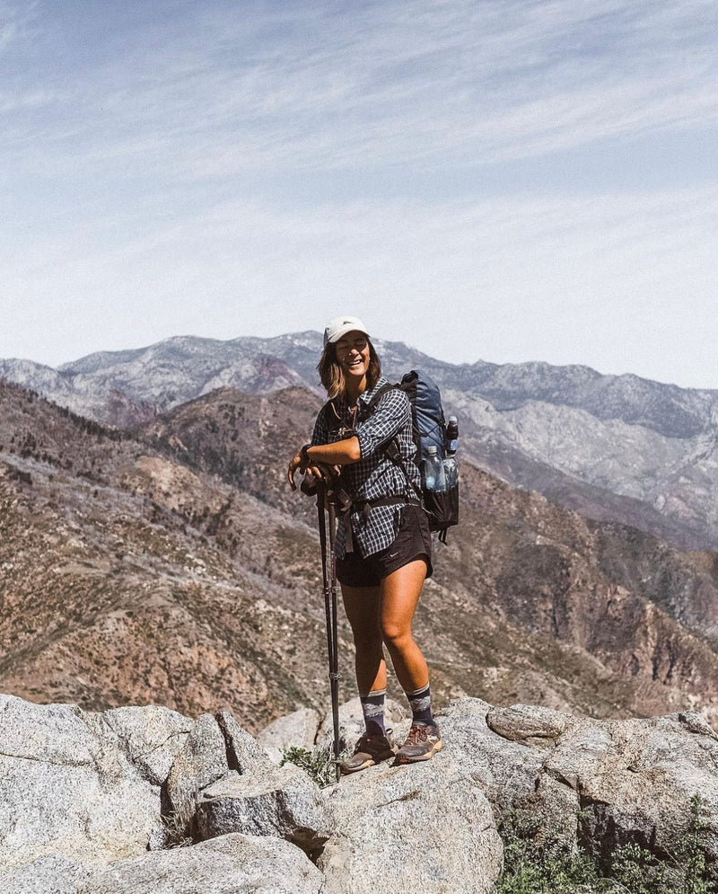 A female hiker smiles at the camera, standing on top of a boulder and resting on her hiking poles.